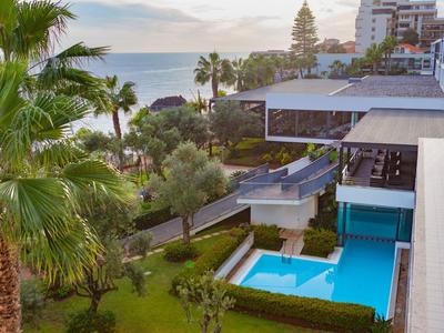 Moderner Hotelbereich mit Pool, grünen Palmen, Weg zur Meeresküste und Gebäuden im Hintergrund.