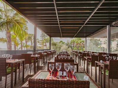 Offenes Restaurant mit Rattanmöbeln, gedeckten Tischen und Blick auf Palmen und Wasser.