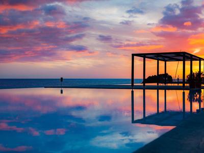Ein ruhiger Pool spiegelt den farbenfrohen Sonnenuntergang mit Wolken und Pavillon am Rand.