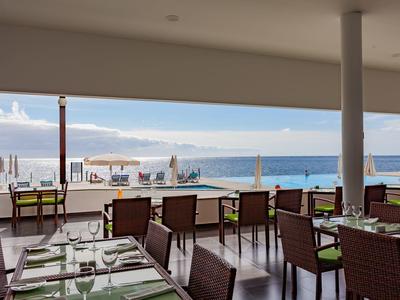 Restaurant mit Tischen und Stühlen, Blick auf Meer, Sonnenschirme und blauen Himmel am Horizont.