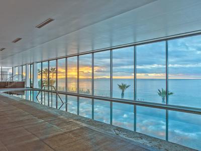 Großes Hallenbad mit Glaswand und Blick auf Meer und Sonnenuntergang, Palmen am Horizont.