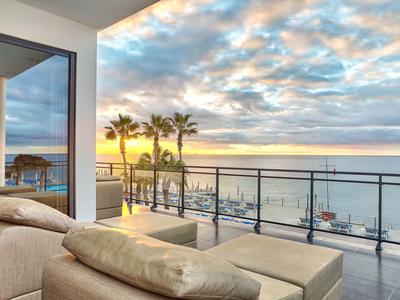 Balkon mit Ledersesseln, Glasgeländer, Palmen und Meer bei Sonnenuntergang mit Wolkenhimmel.