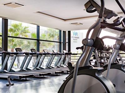 Heller Fitnessraum mit Laufbändern und Crosstrainern vor großen Fenstern und Pflanzen draußen.