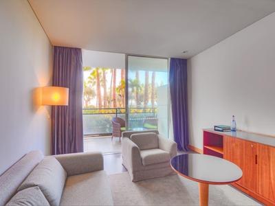 Helles Wohnzimmer mit Sofa, Sessel, Tisch, Kommode und großem Balkon mit Glasgeländer.