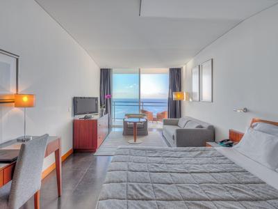 Helles Hotelzimmer mit großem Bett, Schreibtisch, Sofa und Meerblick durch große Fenster.