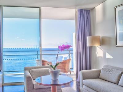 Helles Wohnzimmer mit grauem Sofa, Sesseln, kleinem Tisch und Meerblick durch große Glasfenster.