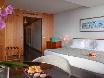 Modernes Hotelzimmer mit großem Bett, Schrank aus Holz, Tisch mit Snacks und lila Blume im Vordergrund.