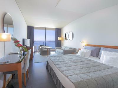 Helles Hotelzimmer mit zwei Einzelbetten, Schreibtisch, Sitzgruppe und großem Fenster mit Meerblick.