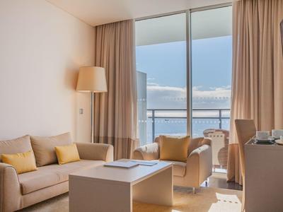 Helles Wohnzimmer mit Sofa, Sessel, weißem Tisch, Vorhängen und Balkon mit Meerblick.