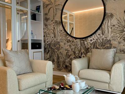 Gemütliches Wohnzimmer mit beige Sesseln, Glas-Couchtisch und rundem Spiegel an Wand mit Pflanzenmuster.