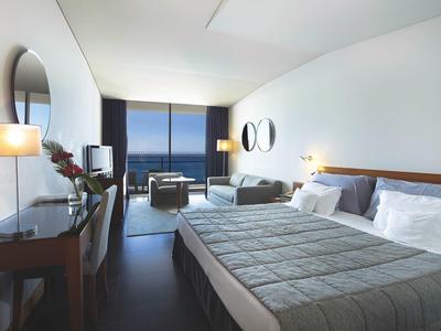 Modernes Hotelzimmer mit großem Bett, Schreibtisch, zwei Sesseln und Meerblick durch bodentiefe Fenster.