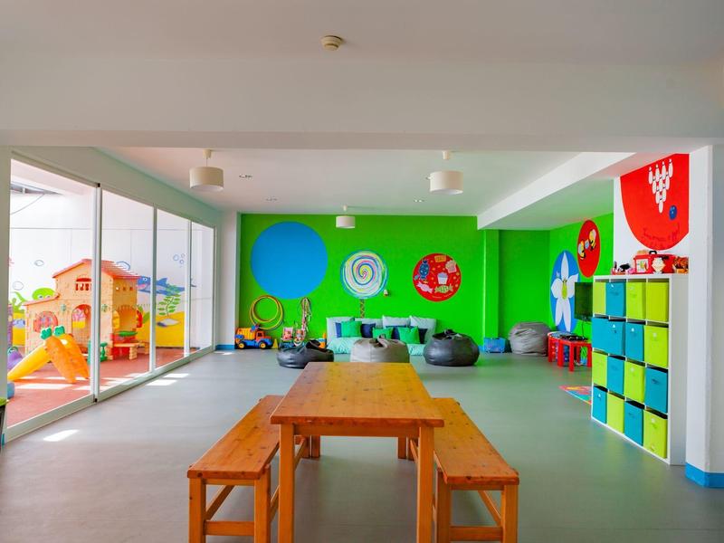 Sala giochi luminosa con pareti verdi, tavoli e sedie in legno, contenitori colorati e giocattoli.