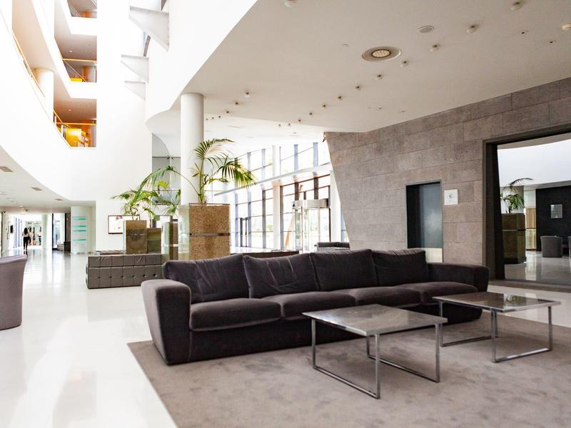 Lobby dell'hotel moderna con divano nero, tavoli in vetro e metallo, pavimento chiaro e luce naturale.