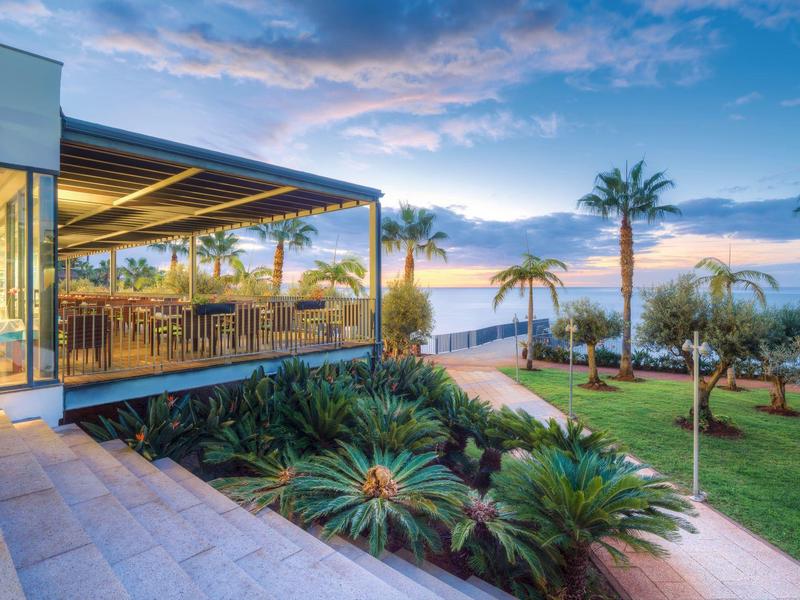 Terrazza moderna con pareti di vetro che si affaccia su giardino tropicale e mare al tramonto.