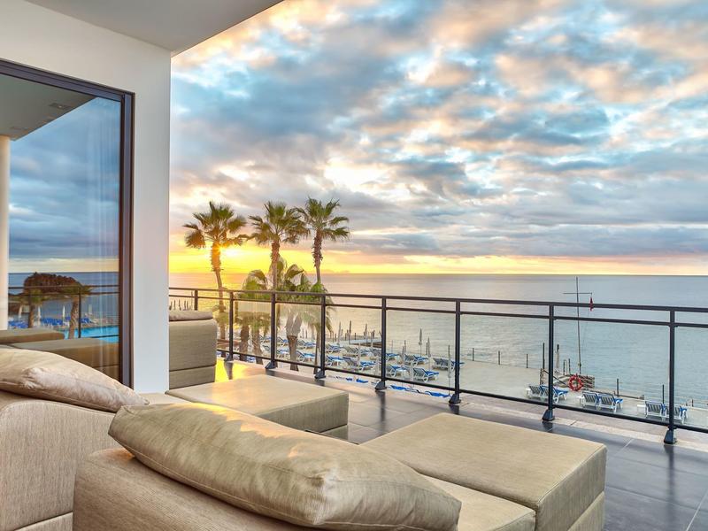 Soggiorno moderno con parete in vetro che si affaccia sul mare con palme al tramonto.