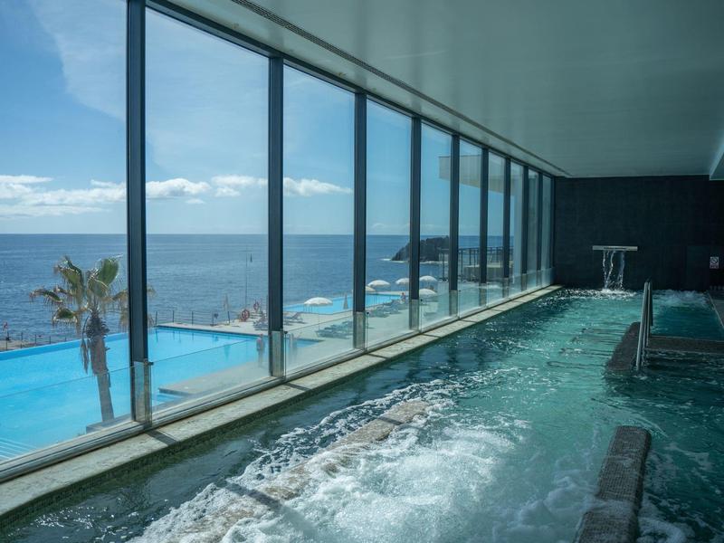 Vasca idromassaggio interna con vista sul mare e piscina esterna sotto cielo sereno.