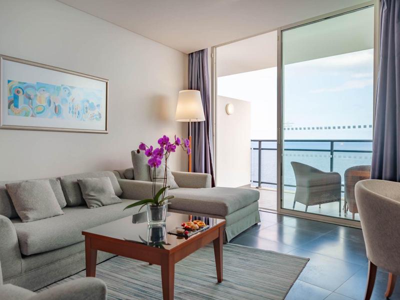 Camera d'hotel moderna con divano, fiori e balcone con vista sul mare.