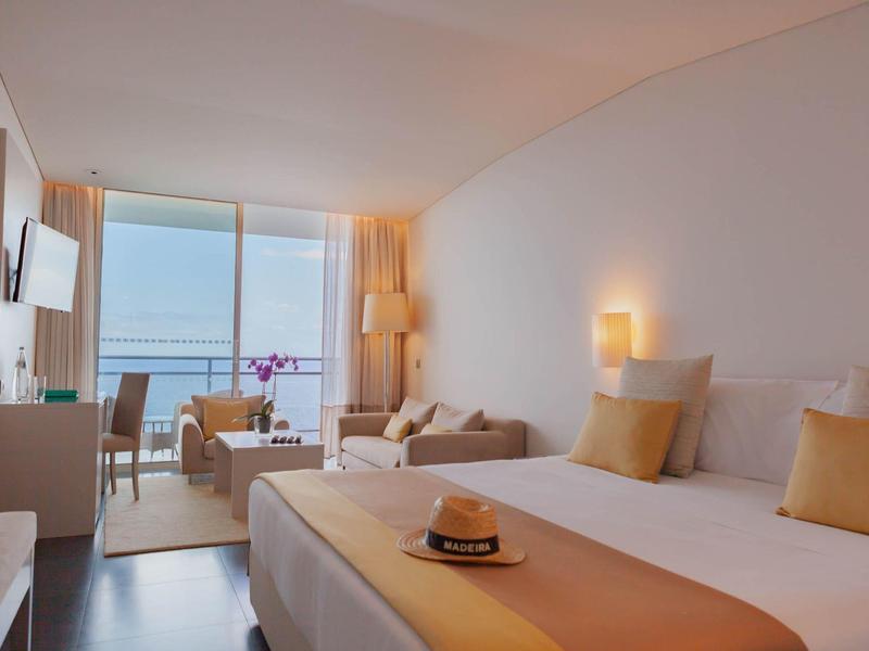 Camera d'hotel luminosa con letto matrimoniale, divano e vista mare attraverso grandi finestre.