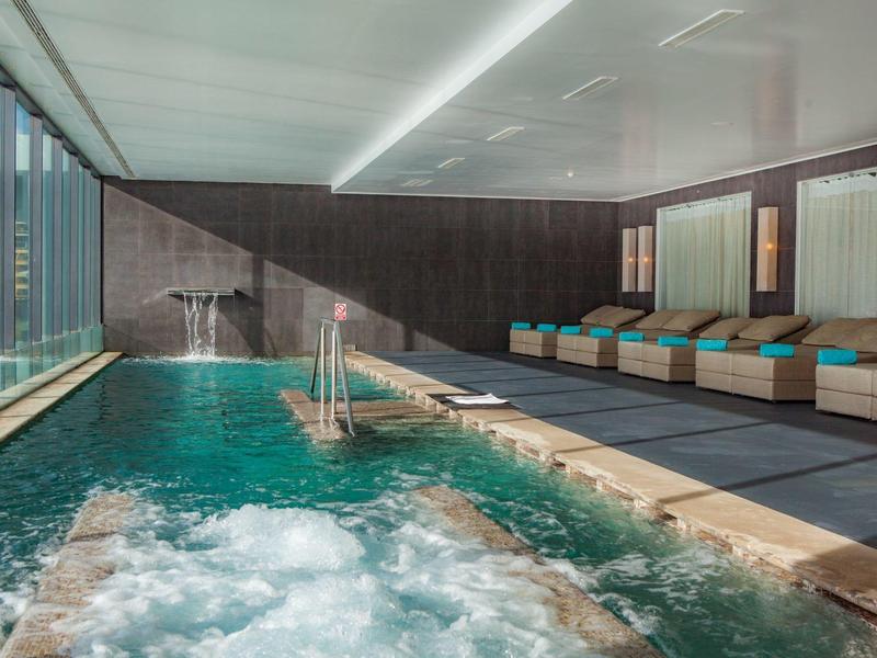 Piscina interna moderna con vasca idromassaggio, lettini e finestre panoramiche.