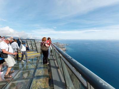 Menschen stehen auf einer gläsernen Aussichtsplattform mit Blick auf den Ozean unter einem bewölkten Himmel.
