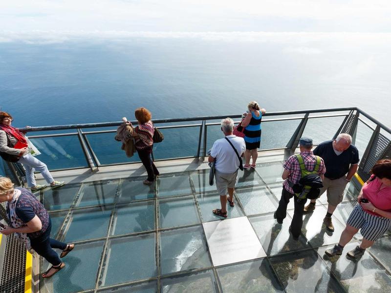 Glasboden-Aussichtsplattform mit mehreren Personen und Blick auf Wolkenmeer und Himmel.