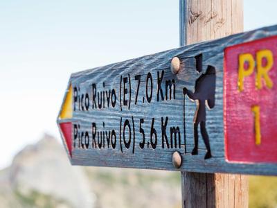 Wanderwegweiser auf Holz mit Entfernungsangaben und Wanderersymbol vor Bergkulisse.