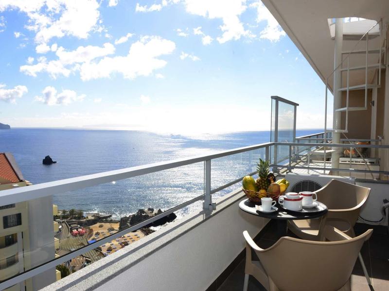Balkon mit Tisch, Stühlen und Obstkorb, Blick auf Meer, Häuser und bewölkten Himmel.