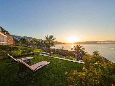 Sonnenuntergang über Wasser mit grüner Wiese, Liegestühlen und Palmen im Vordergrund.