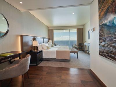 Modernes Schlafzimmer mit großem Bett, Schreibtisch, Sessel und Meerblick durch großes Fenster.