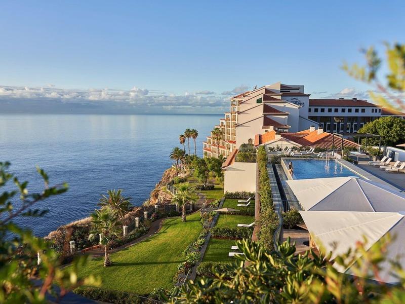 Hotelgebäude auf Klippe mit Blick auf ruhiges, blaues Meer und grüne Gartenanlagen bei klarem Himmel.