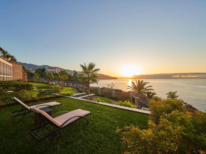 Sonnenuntergang über Wasser mit grüner Wiese, Liegestühlen und Palmen im Vordergrund.