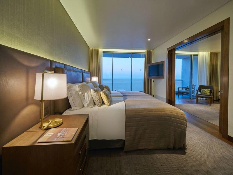 Modern eingerichtetes Schlafzimmer mit großem Bett, Nachttisch, Lampe und Meerblick durch Panoramafenster.