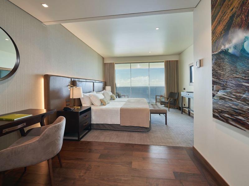 Modernes Schlafzimmer mit großem Bett, Schreibtisch, Sessel und Meerblick durch großes Fenster.