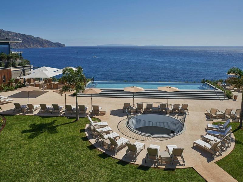 Blick auf einen Poolbereich mit Sonnenliegen und Palmen, dahinter das blaue Meer und ein Hügel im Hintergrund.