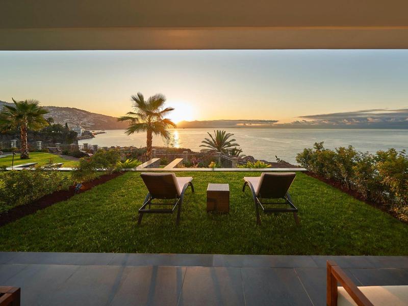 Sonnenuntergang über Meer, zwei Stühle auf grünem Rasen, Palmen und Berge im Hintergrund.