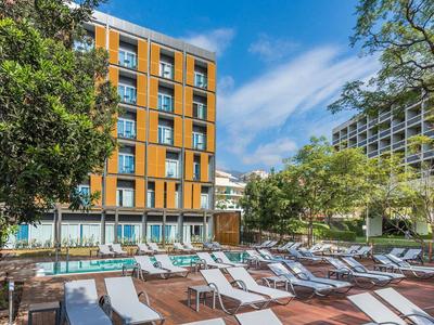 Freiluftpoolbereich mit Liegestühlen, umgeben von Bäumen und einem hohen Gebäude bei blauem Himmel.