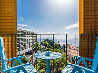 Balkon met blauwe tafel en stoelen, uitzicht op gebouwen en zee in fel zonlicht.