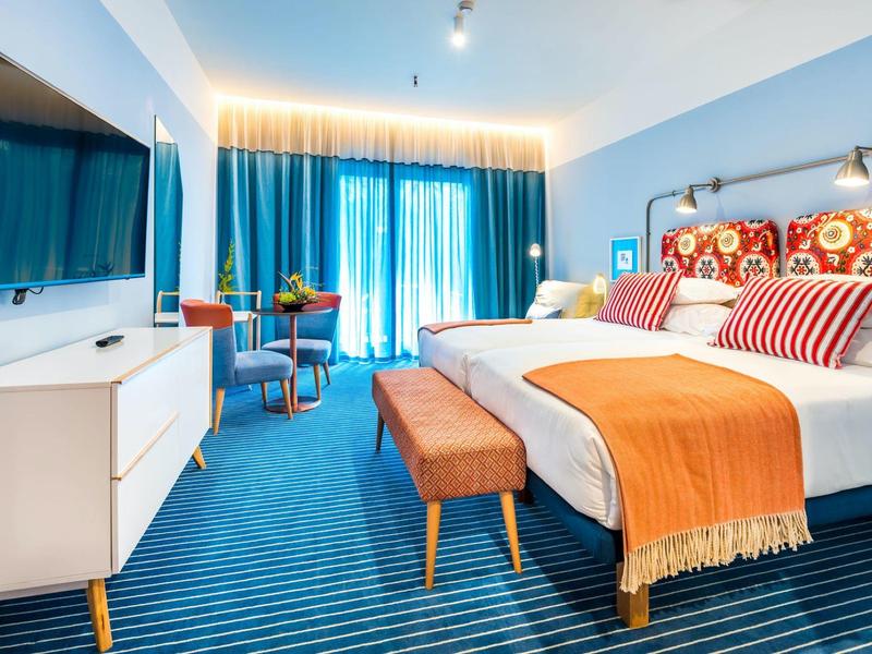 Ein helles Hotelzimmer mit blauem Teppich, Doppelbett, orangefarbenen Akzenten und großem Fenster.