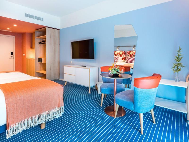 Modernes Hotelzimmer mit blauem Teppich, orangen Akzenten und minimalistischem Mobiliar.