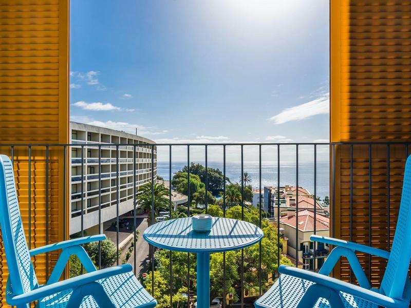 Balkon mit blauem Tisch und Stühlen, Blick auf Stadt, Bäume und hellen Himmel bei Sonnenschein.