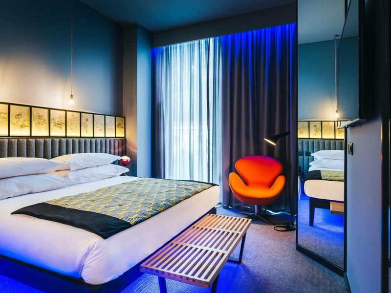 Modernes Hotelzimmer mit großem Bett, leuchtender blauer und roter Akzentbeleuchtung und breiten Vorhängen.