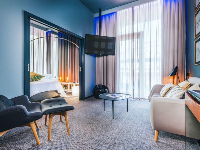 Modernes Wohnzimmer mit großen Fenstern, Vorhängen, zwei Sesseln, Sofa, Spiegel und Flachbildschirm.