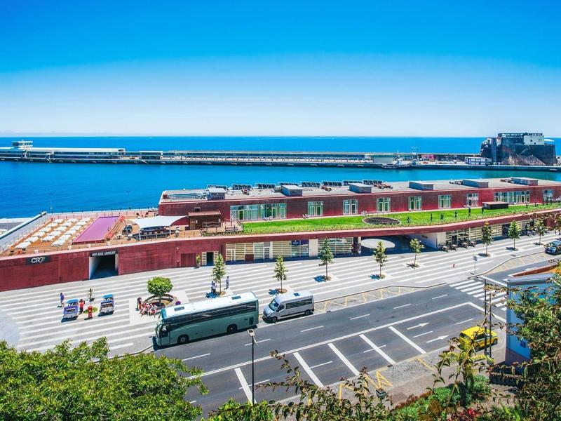 Hafenanlage mit langem, rotem Flachdachgebäude, mehreren Bussen und blauem Meer im Hintergrund.