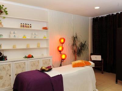 Behandlungsraum mit Massageliege, lila Decke, orange Kissen, Regal und warmem Stehlampenlicht.