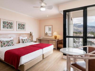 Bright hotel room with two beds, table and chairs, balcony overlooking mountains.