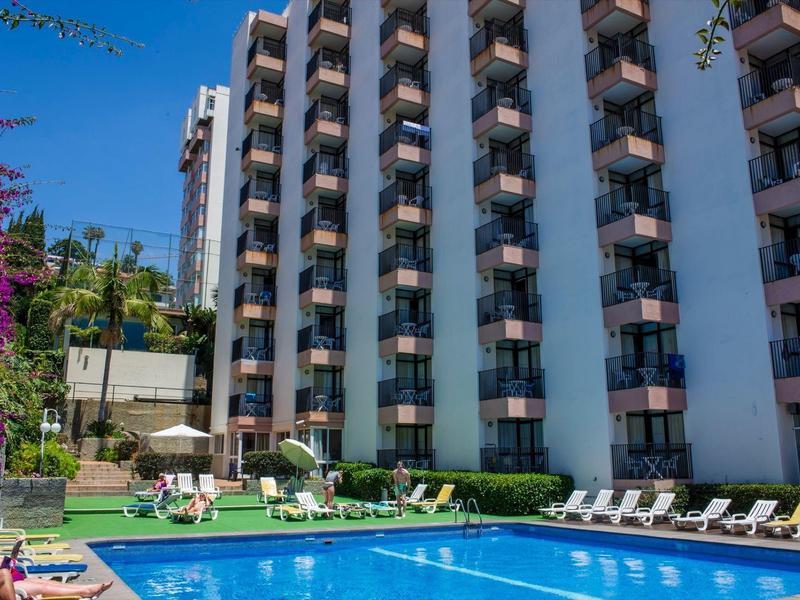 Mehrstöckiges Hotel mit Balkonen, großem blauen Pool, Liegestühlen und Sonnenschein im Außenbereich.