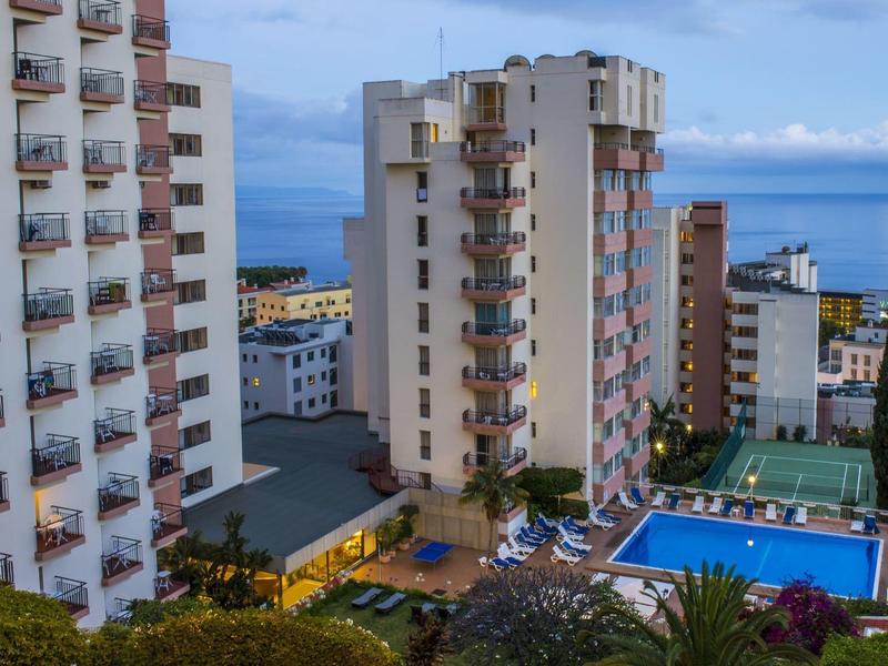 Mehrere hohe Wohnhäuser mit Balkonen, ein Pool und Meer im Hintergrund bei klarem Himmel.