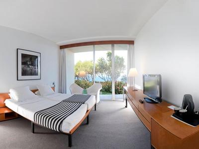 Camera d'hotel moderna con due letti, scrivania, TV e grande finestra con vista su alberi verdi.