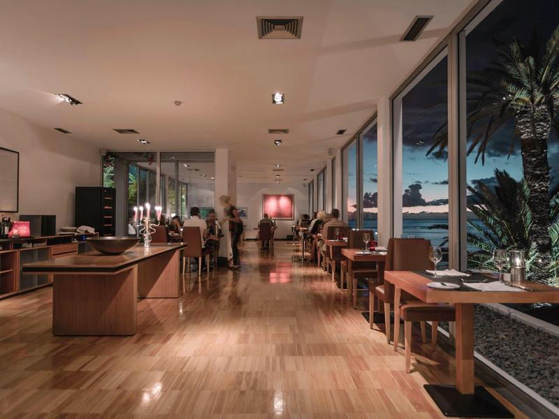 Area ristorante moderna dell'hotel con pavimento in legno, file di tavoli e grandi vetrate al crepuscolo.