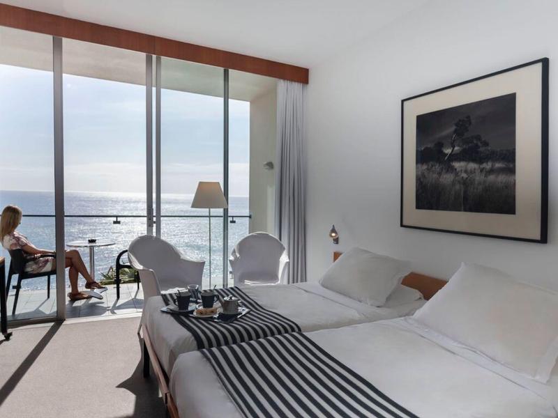 Camera d'hotel luminosa con vista mare e balcone dove una donna è seduta.